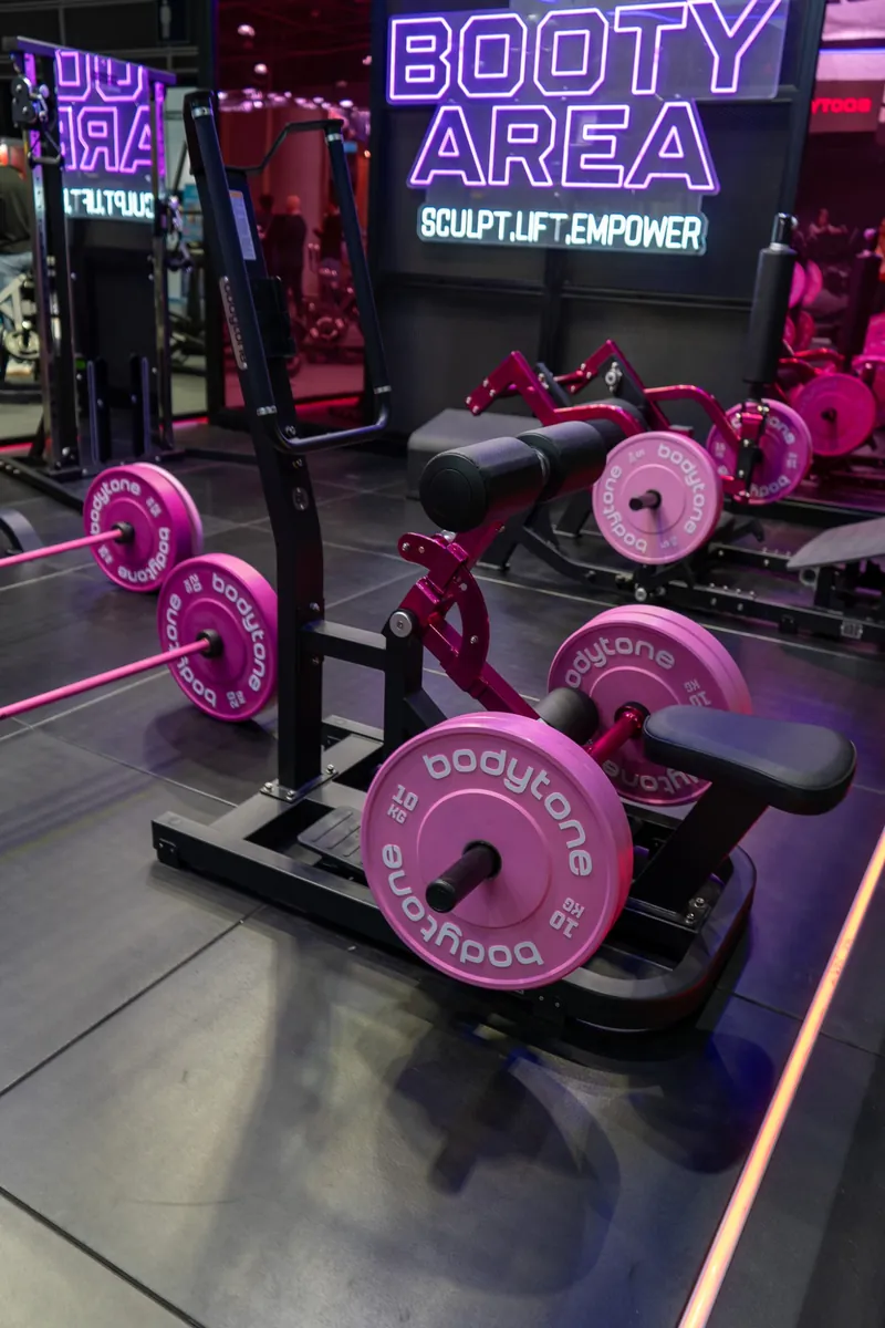 Der Neon-Schriftzug „Booty Area — Sculpt. Lift. Empower.“ über den Geräten der Zone.