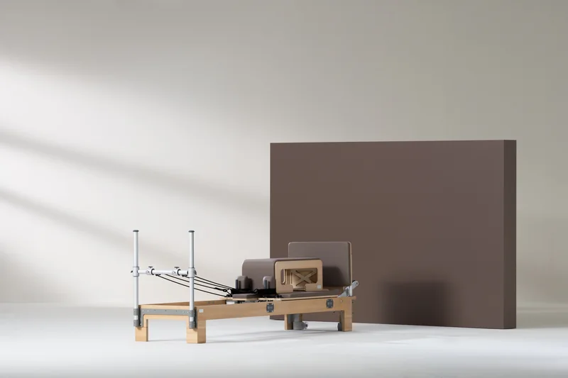 Ein BASI Systems Reformer mit Holzrahmen, gepolstertem Wagen und aufgestellten Risern, vor hellem Studiohintergrund fotografiert.